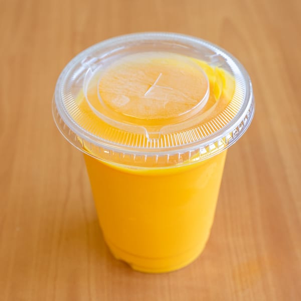 A vibrant orange drink in a clear plastic cup with a lid, on a light wooden surface.